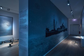 Ein blau beleuchteter Raum mit steinernen Elementen auf dem Boden und einer schwarz-weißen Wandtapete mit einem Schiff.