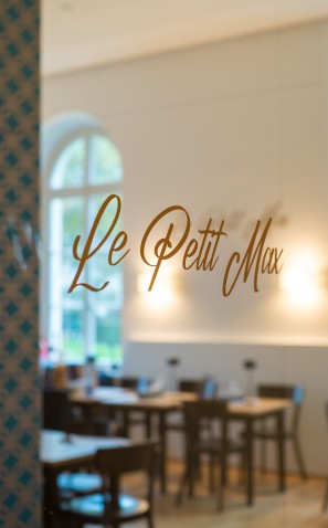 Eine Glastür mit Schriftzug Le Petit Max, im Hintergrund das Café mit Tischen und Stühlen
