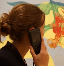 Foto: Eine junge Frau im Profil, die sich einen Audioguide an ihr Ohr hält, vor der Ancolie von Max Ernst